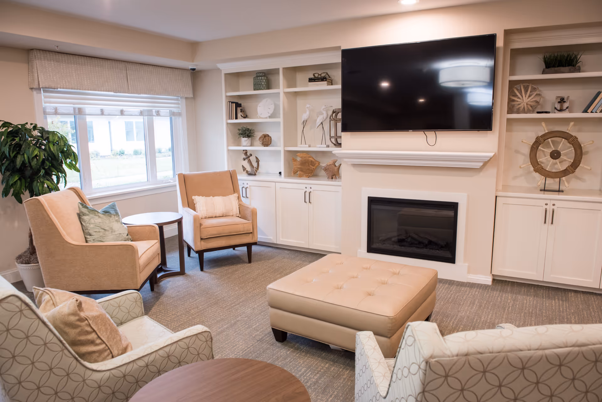 Comfortable seating area with armchairs, an ottoman, built-in shelves, a wall-mounted TV and a fireplace in a senior living common room.