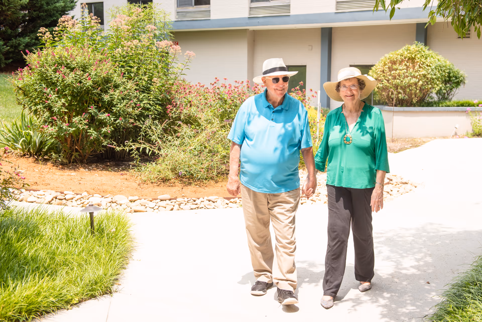 An elderly couple holding hands and walking on a sunny paved pathway surrounded by greenery and bushes outside a building.