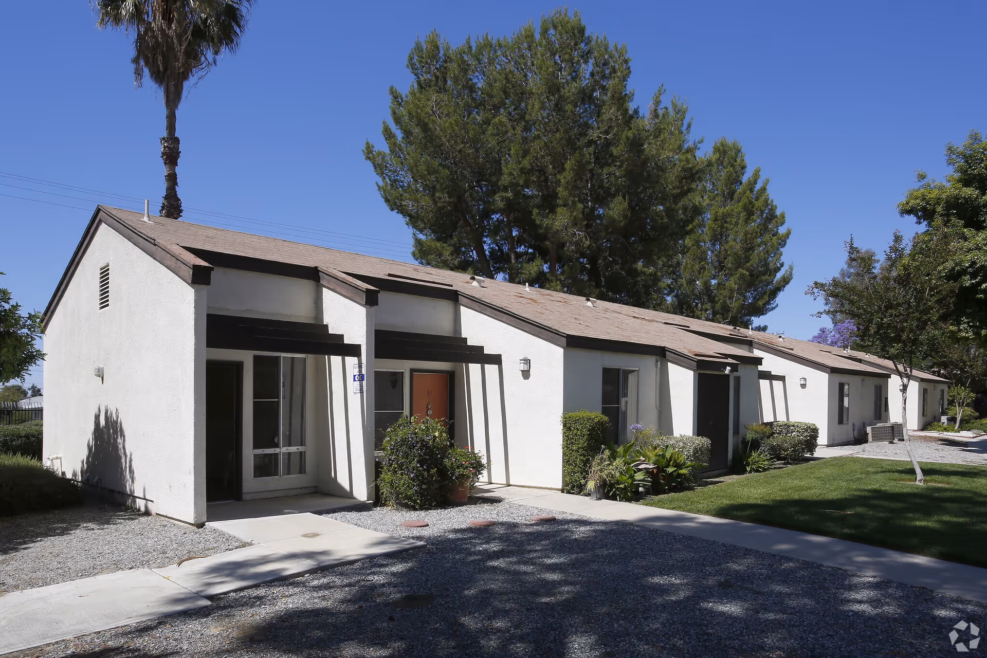 Exterior view of a single-story residential building with white walls and brown roof shingles, surrounded by trees and greenery under a clear blue sky.
