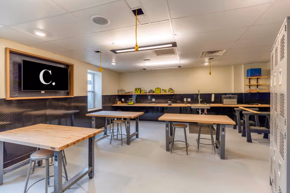 A well-lit workshop room with several wooden workbenches and metal stools. The walls have a black textured panel halfway up, and there are tools and equipment on shelves along the back wall. A large screen with the letter 'C.' is mounted on the left wall. The ceiling has fluorescent lights and yellow hanging cords. Lockers are visible on the right side of the room.