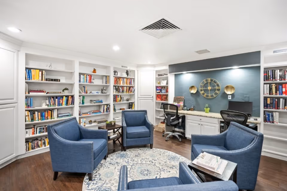 A cozy interior room with four blue upholstered armchairs arranged around a circular patterned rug. Behind the chairs are built-in white bookshelves filled with books and decorative items. To the right, there is a workspace with two office chairs, a granite countertop, a computer monitor, and a wall clock with gold accents on a dark blue wall.