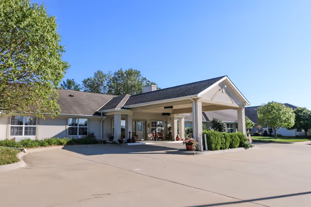 Exterior view of a single-story senior living facility building with a covered entrance, surrounded by trees and shrubs under a clear blue sky.