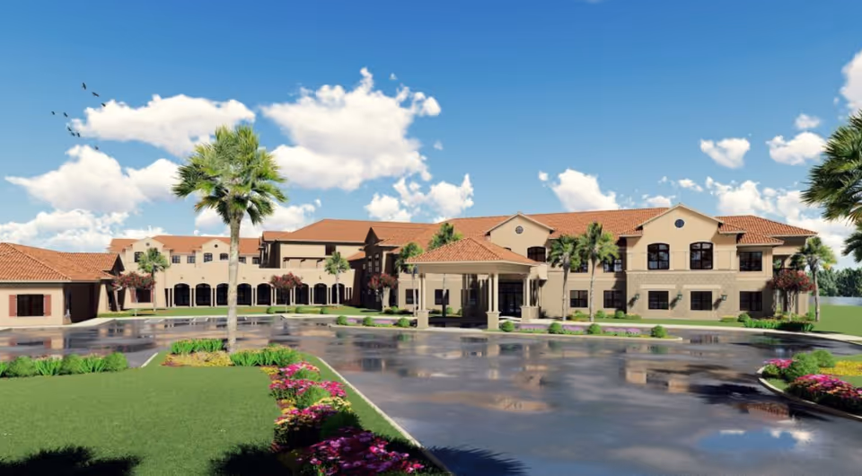 Exterior view of a large senior living facility building with a red-tiled roof, beige walls, palm trees, and landscaped flower beds under a blue sky with scattered clouds.