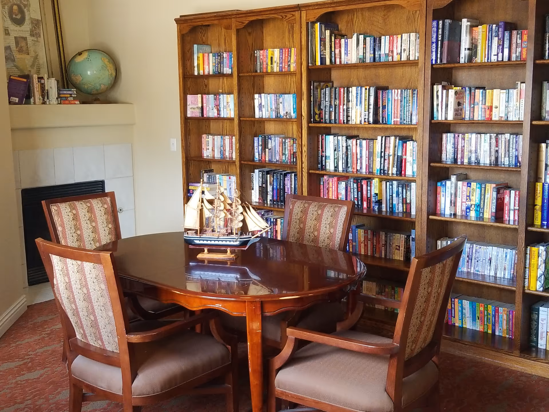 A cozy library-style room with a round wooden table, four upholstered chairs, a model ship on the table, and tall bookshelves filled with books.