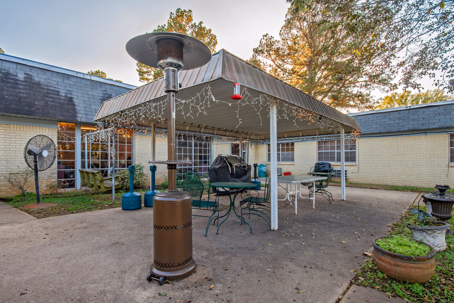 Outdoor patio area at The Blossoms at Stamps Rehab & Nursing Center with a covered seating area featuring metal tables and chairs, a standing patio heater, a large fan, and potted plants. The building with yellow brick walls and multiple windows surrounds the patio, and trees are visible in the background.