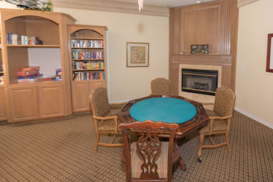 A cozy room with a round wooden table covered with a green felt top surrounded by four upholstered chairs. Behind the table is a wooden fireplace with a framed picture above it. To the left, there are wooden bookshelves filled with books and board games. The room has beige walls and patterned carpet flooring.