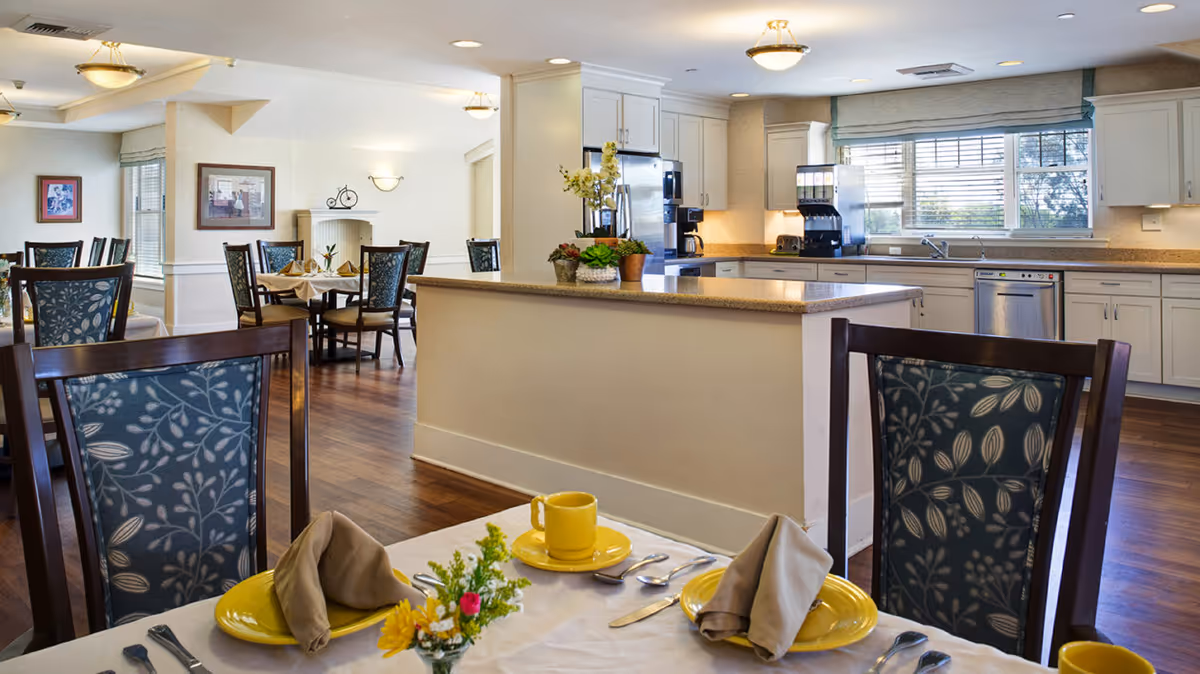 A bright and clean dining area with several tables and chairs, set with yellow plates, beige napkins, and silverware. In the background, there is a kitchen area with white cabinets, a countertop island, a refrigerator, a coffee maker, and a beverage dispenser. The room has wooden flooring, large windows with blinds, and framed pictures on the walls.