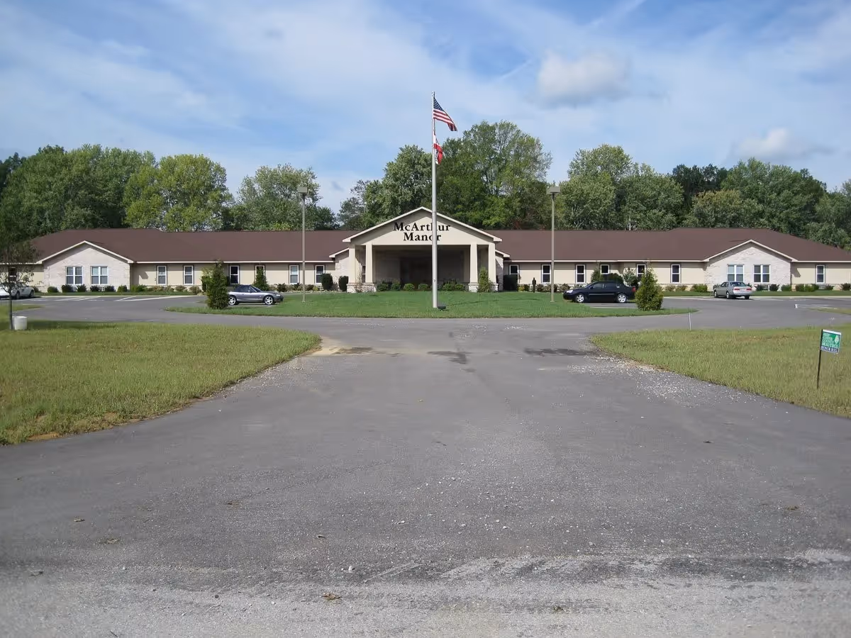 Front exterior of McArthur Manor assisted living building with a central covered entrance, flagpole, lawn, and parking lot.