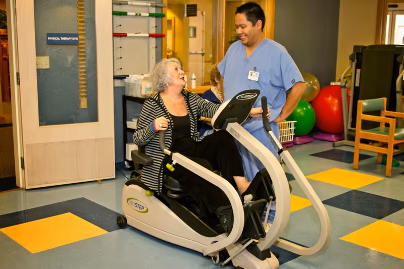 An elderly woman using a recumbent exercise bike in a physical therapy gym, smiling and interacting with a male healthcare professional in blue scrubs who is standing beside her.