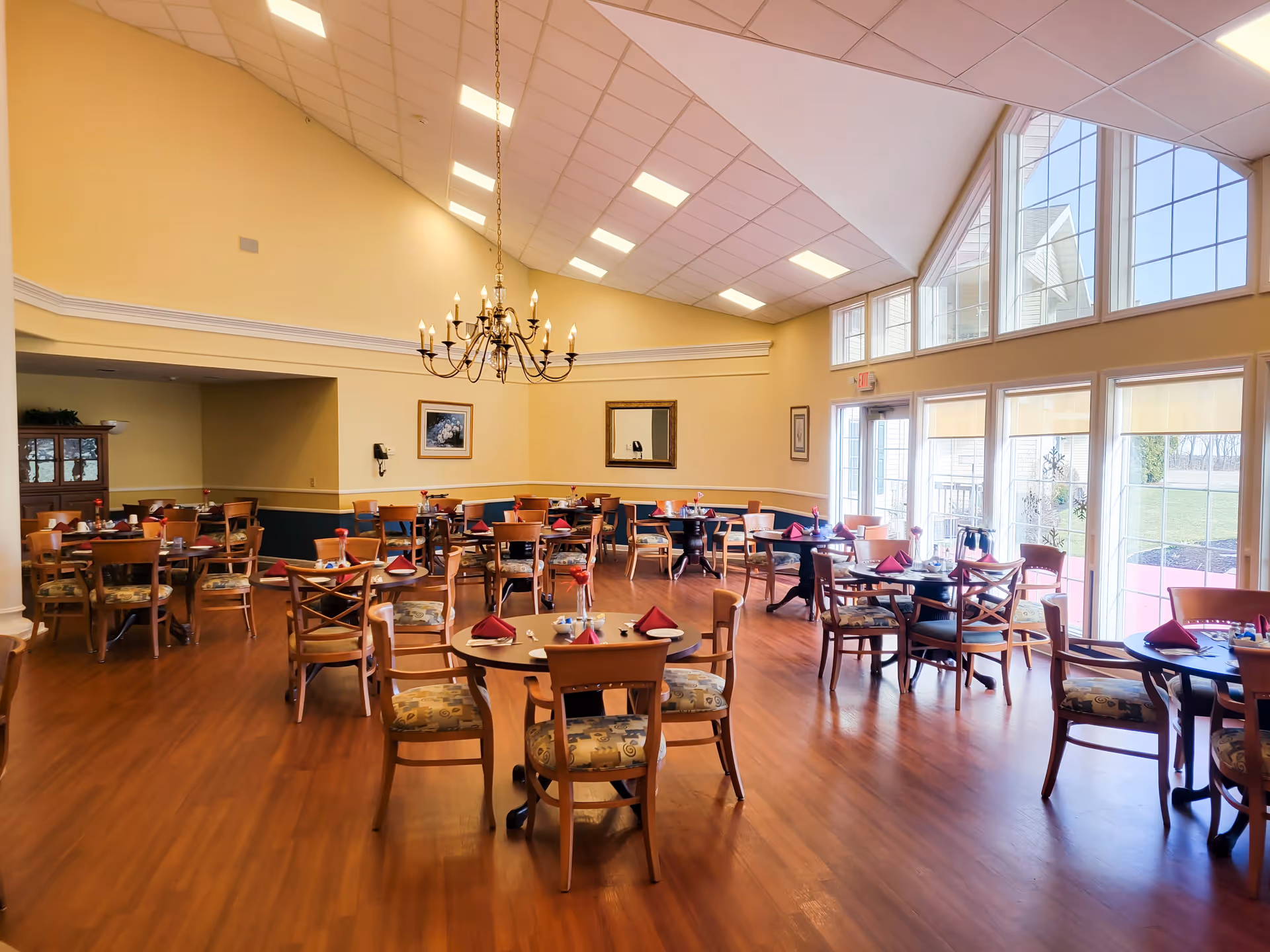 A spacious dining room with multiple round wooden tables and chairs arranged neatly. Each table is set with red napkins, plates, and silverware. The room has large windows on one side allowing natural light to fill the space, and a chandelier hanging from the ceiling. The walls are painted yellow with white trim, and there is a wooden cabinet and framed artwork on the walls.