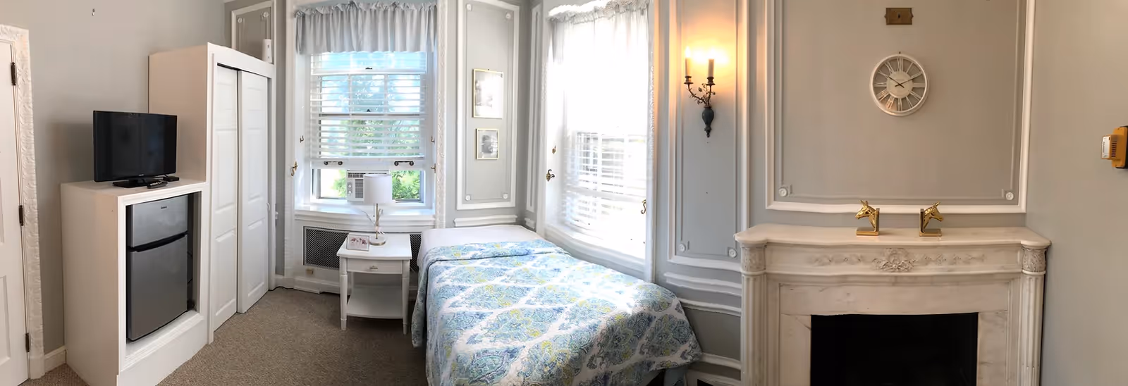 A small, cozy bedroom with a single bed covered in a blue and green patterned quilt. Next to the bed is a white nightstand with a lamp. There is a window with white blinds and an air conditioning unit. A white wardrobe and a small refrigerator with a television on top are positioned against the wall. The room features a decorative white fireplace with two gold horse head bookends and a round wall clock above it. The walls are painted light gray with white trim and molding, and a wall sconce light is mounted near the fireplace.