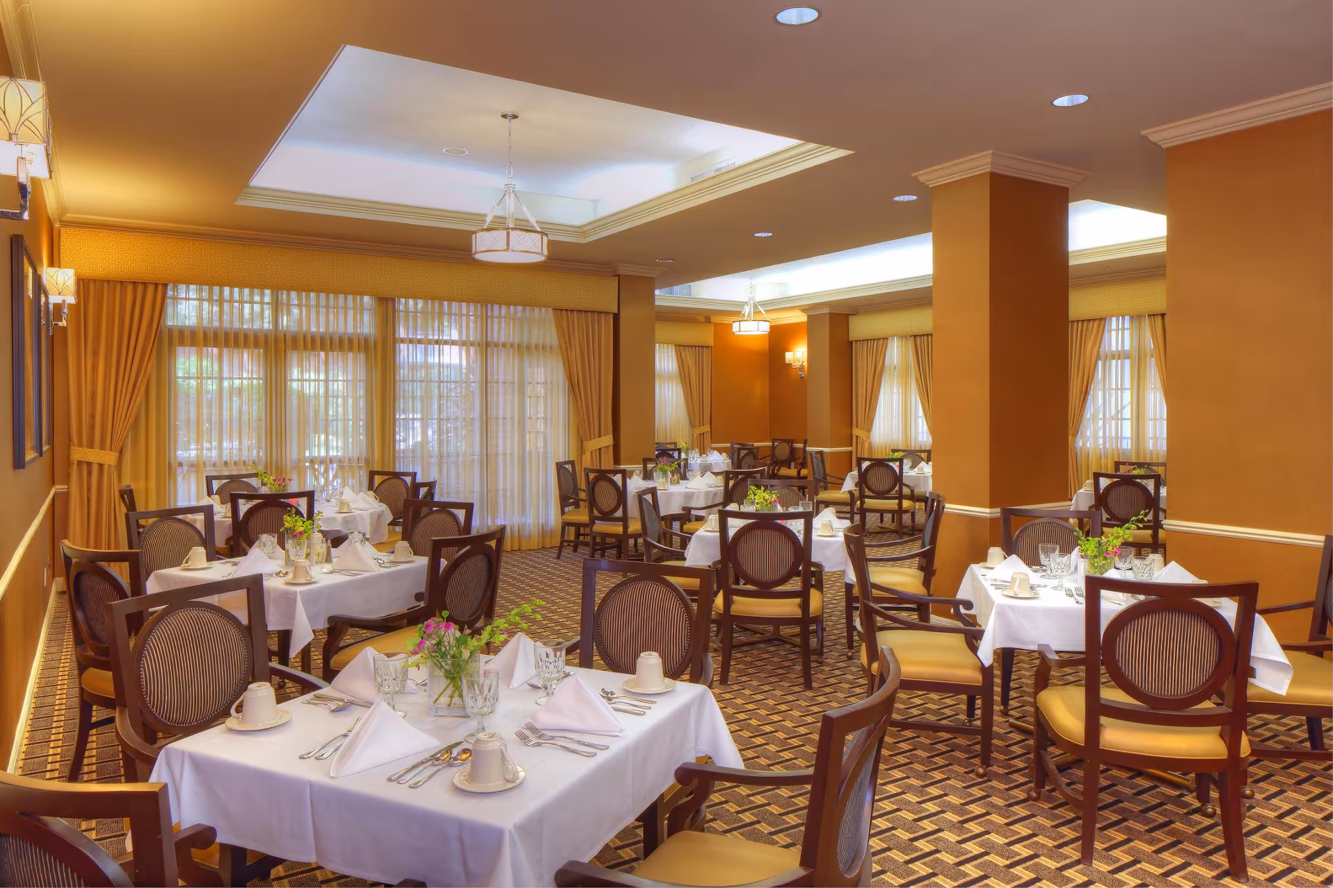 A warmly lit dining room with multiple tables covered in white tablecloths, each set with folded napkins, cups, glasses, and silverware. The room features patterned carpet, wooden chairs with cushioned seats, large windows with sheer curtains and gold drapes, and ceiling light fixtures.