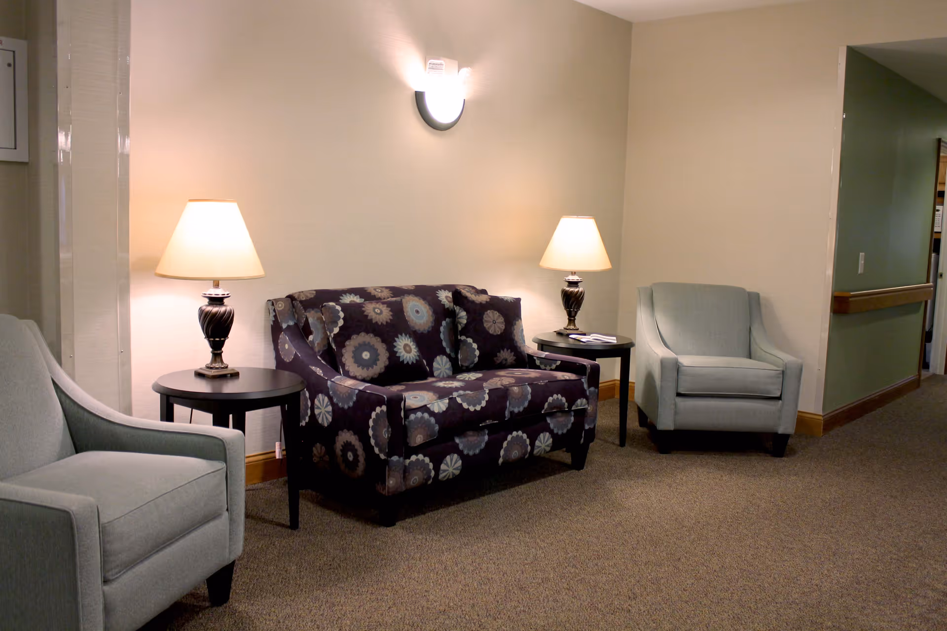 A cozy seating area in a hallway with a patterned loveseat, two light blue armchairs, two side tables each with a lamp, and a wall-mounted light fixture above the loveseat.