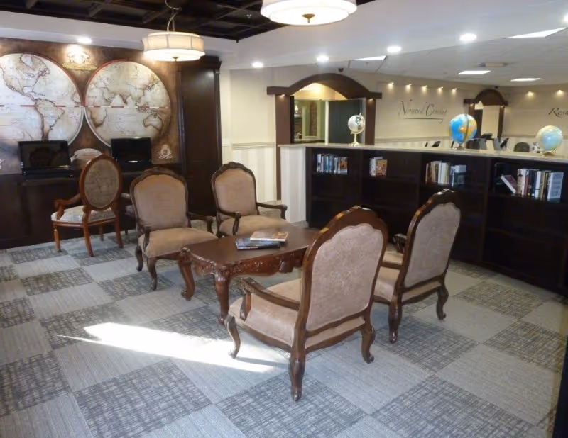 A cozy sitting area with four upholstered wooden armchairs arranged around a wooden coffee table. Behind the seating area is a dark wood bookshelf with books and globes on top. The wall features two large vintage-style world maps and two computer stations with chairs. The room has a patterned carpet and a ceiling with recessed lighting and a hanging light fixture.