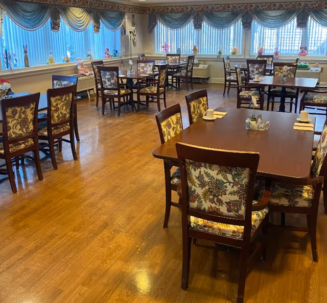 A dining room with multiple wooden tables and chairs featuring floral upholstery. The room has large windows with blue vertical blinds and valances, decorated with small figurines on the windowsills. The floor is wooden, and the tables are set with cups, napkins, and condiment holders.