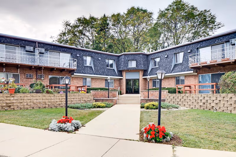 Exterior view of Whitcomb Square Apartments showing a two-story brick building with multiple windows and balconies. There is a concrete walkway leading to the main entrance, flanked by two black lamp posts with flower beds at their bases. The building is surrounded by green grass, shrubs, and trees in the background.