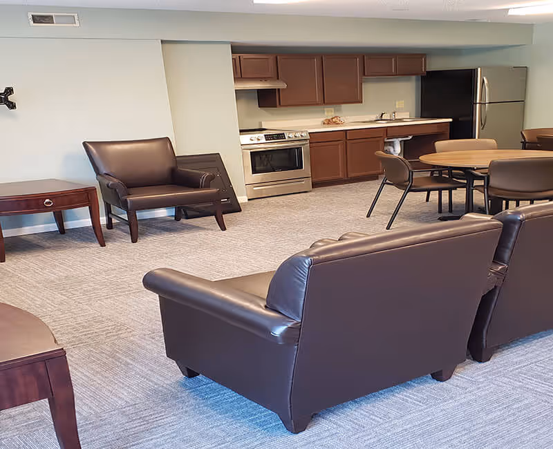 A common area in a senior living facility featuring brown leather armchairs and sofas, a round wooden table with chairs, and a kitchen area with wooden cabinets, a stainless steel stove, and a refrigerator. The room has light-colored walls and carpeted flooring.
