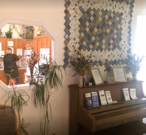 Interior common area with a piano displaying brochures and framed items, a large quilt on the wall, plants, and a view into a kitchen where a staff member is working.