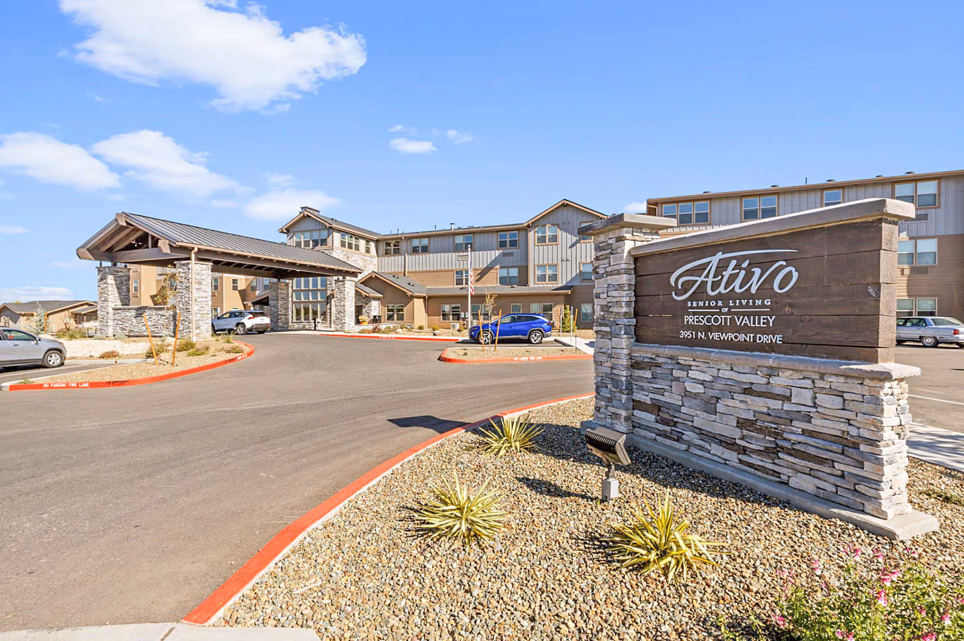 Exterior view of Ativo Senior Living of Prescott Valley showing the entrance driveway, a large stone and wood sign with the facility name and address, and a multi-story building under a partly cloudy sky.