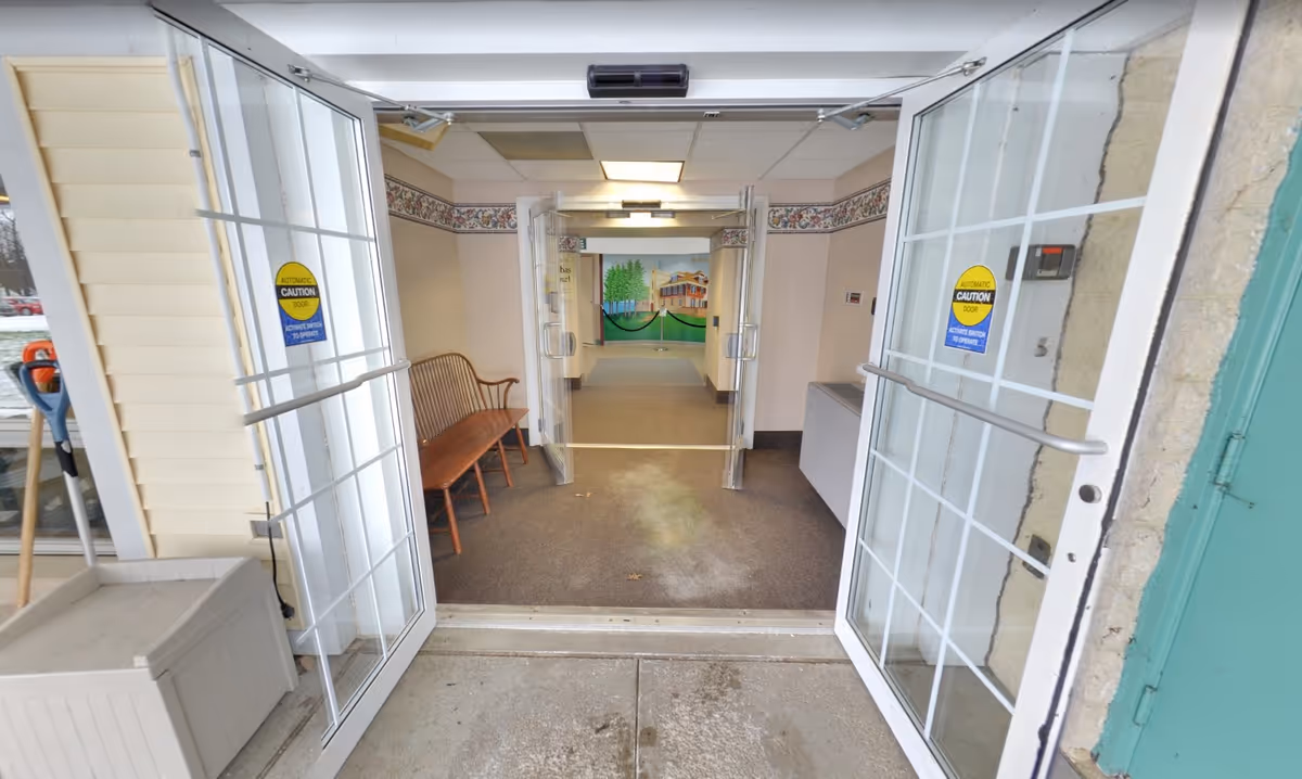 View through open glass double doors into an entryway of a building. Inside, there is a wooden bench along the left wall and a mural of a house and trees on the far wall. The floor is carpeted and the walls have a floral border near the ceiling. There are automatic door caution signs on the glass doors.