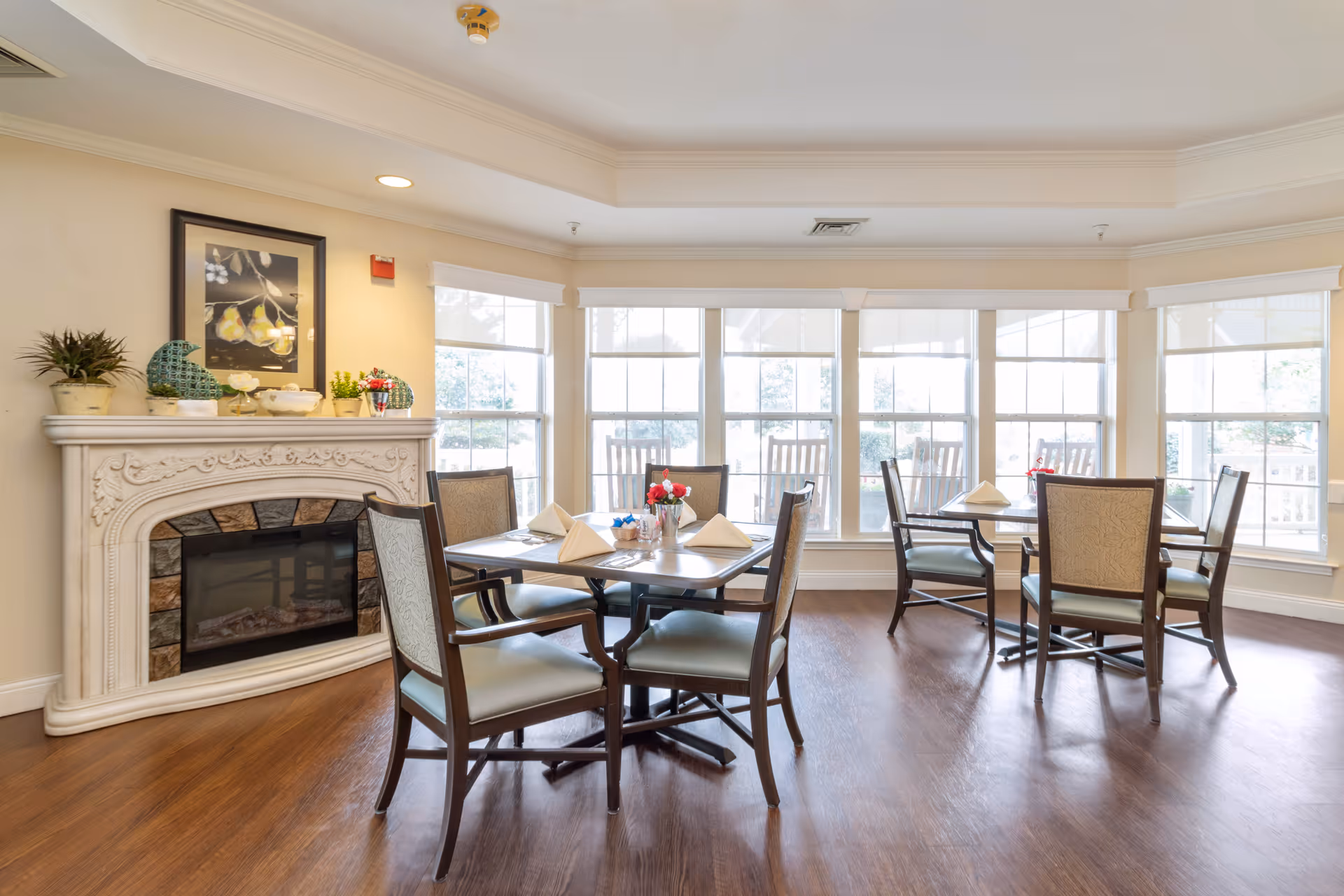 A bright dining room with wooden floors, several tables set with napkins and small flower arrangements, surrounded by cushioned chairs. Large windows let in natural light, and there is a decorative white fireplace with plants and artwork above it on the left side.