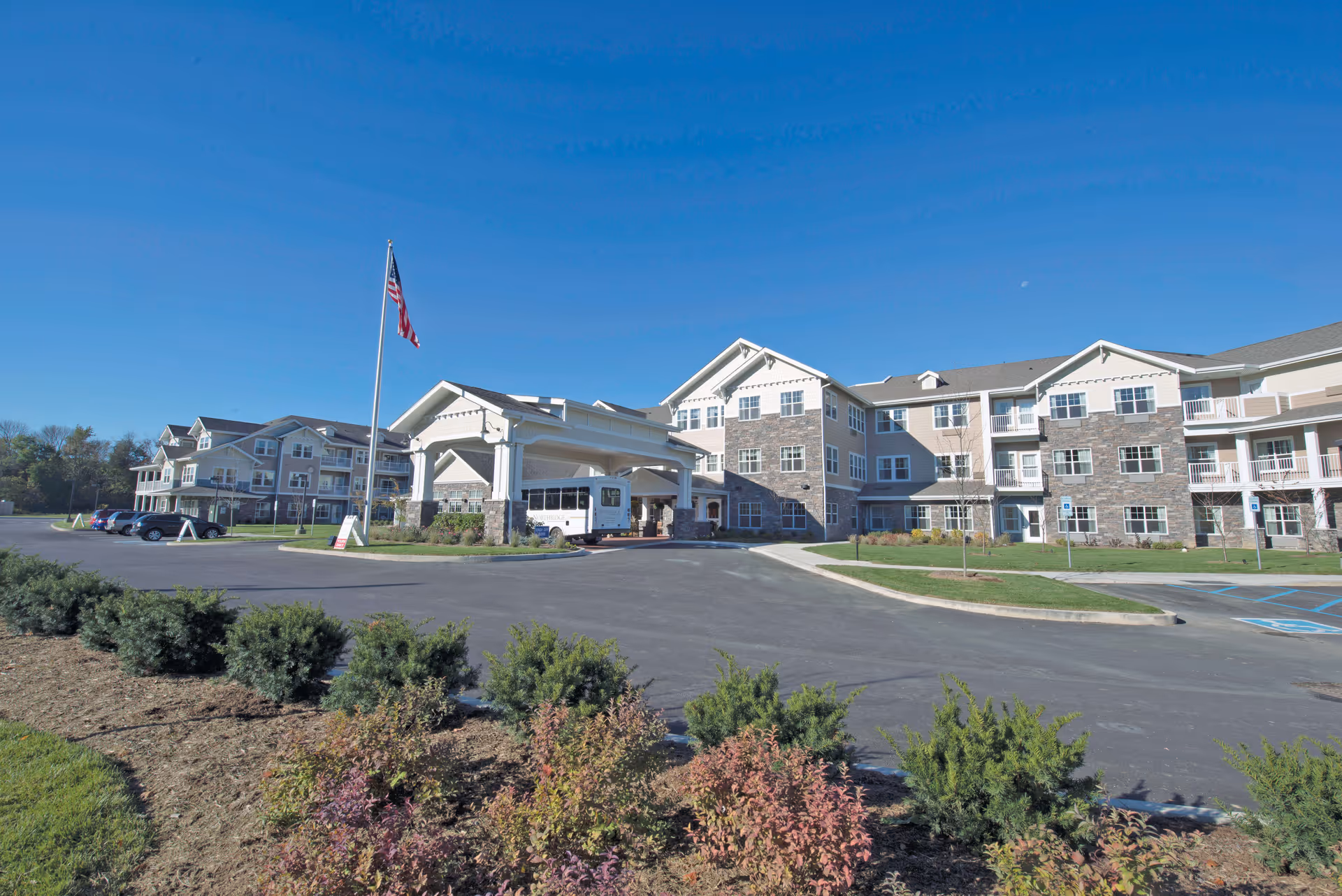 Exterior view of Northridge Gracious Retirement Living facility showing a large multi-story building with stone and siding facade, a covered entrance with a white canopy, an American flag on a flagpole, parked cars, and landscaped bushes in the foreground under a clear blue sky.