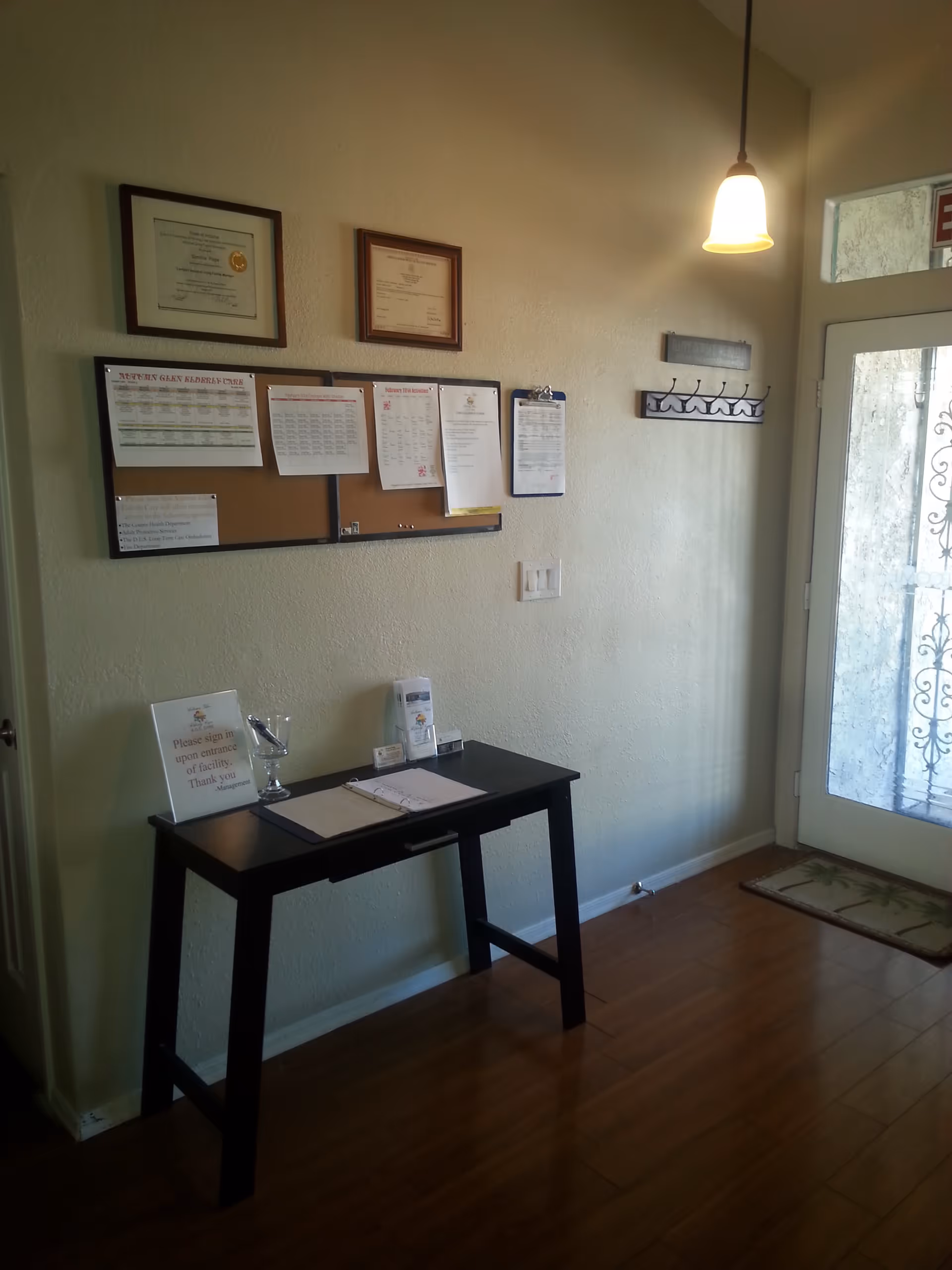 Interior view of an entrance area in Autumn Glen Elderly Care facility showing a small black table with a sign-in book, a glass bowl, and informational pamphlets. Above the table, there is a bulletin board with various notices and two framed certificates on the wall. A hanging light fixture illuminates the area near a glass door with a decorative metal security screen and a floor mat.