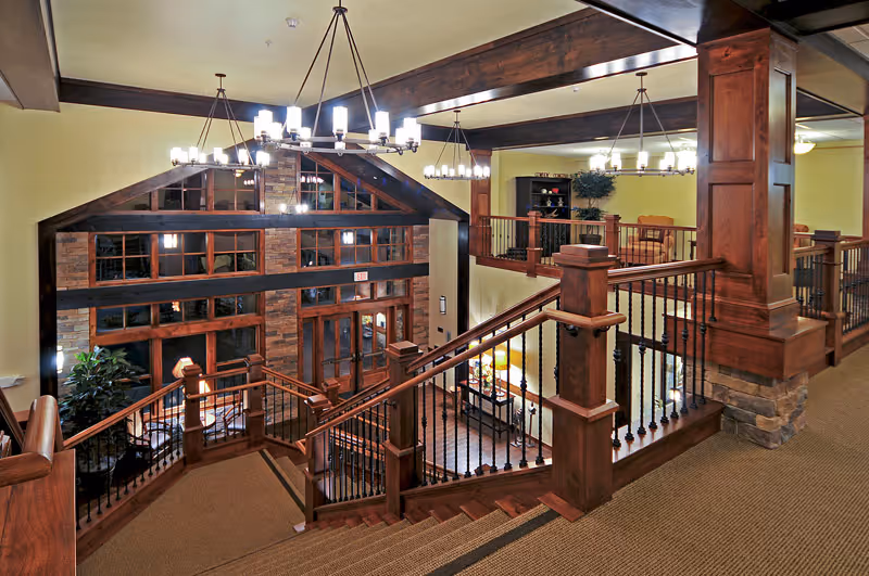 Interior view of a senior living facility featuring a wooden staircase with black metal railings, warm lighting from chandeliers, and large windows with wooden frames. The area includes a seating space with chairs and tables, and the walls are painted in a soft yellow color.