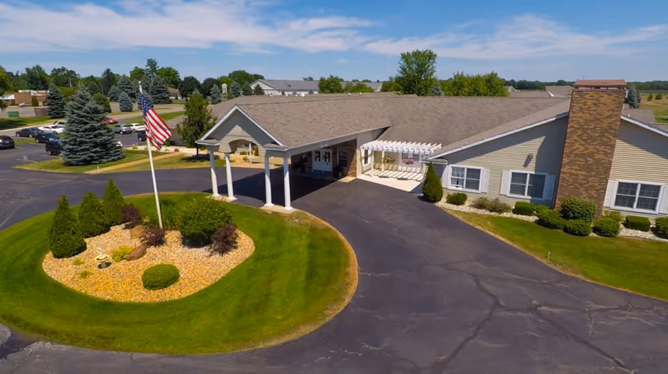 Aerial view of the front entrance of Spring Arbor Assisted Living facility showing a circular landscaped area with an American flag, surrounded by a paved driveway and parking lot. The building has a covered entrance with white columns and a pergola, beige siding, and a brick chimney. Trees and greenery are visible in the background under a partly cloudy sky.