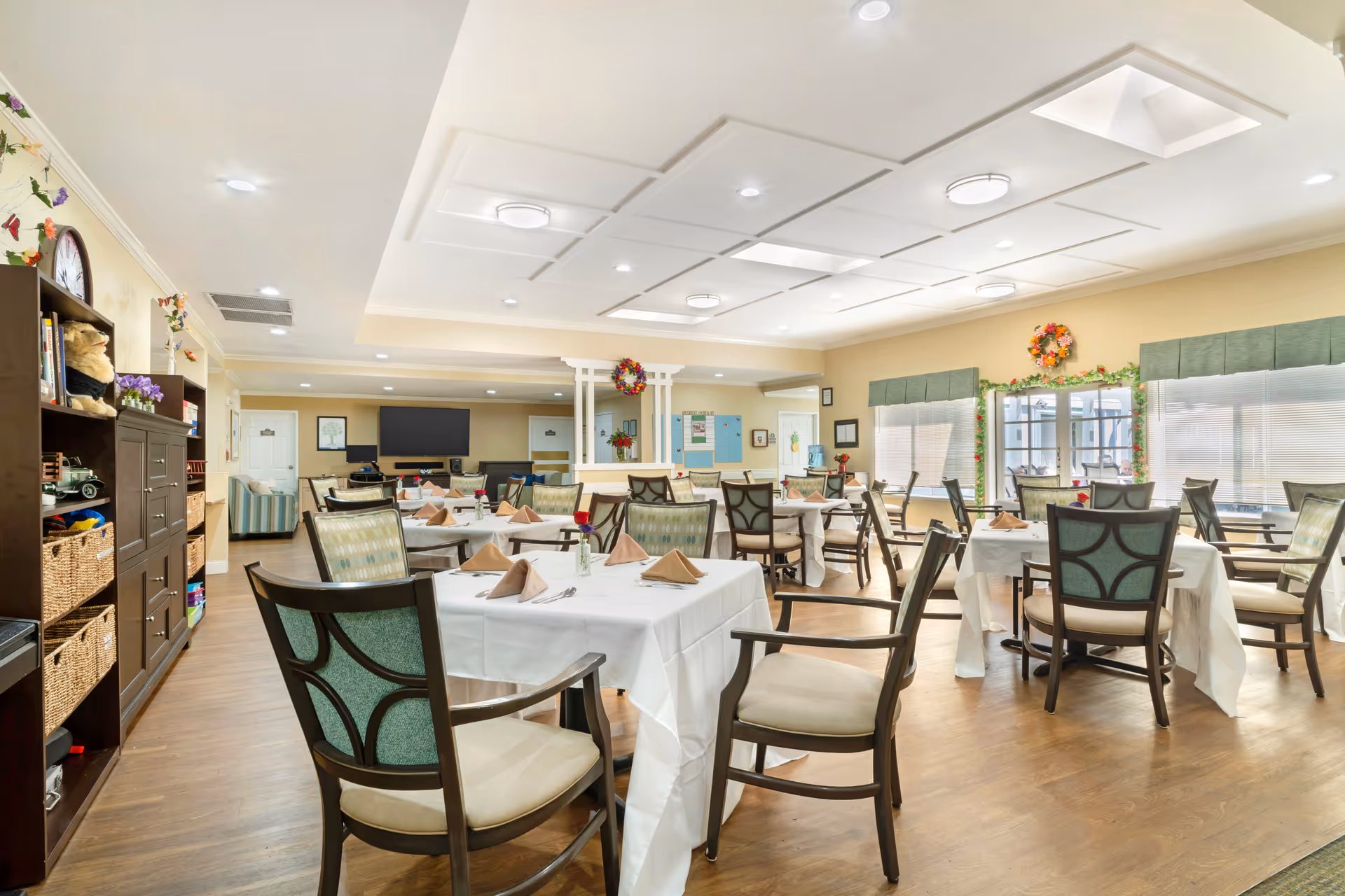 Bright communal dining room with multiple set tables and chairs, windows, and a television at the far wall.