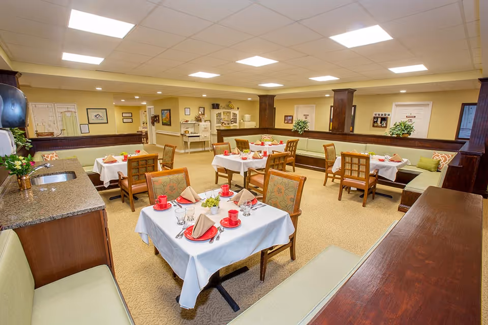 A dining room in a senior living facility with several tables covered with white tablecloths, each set with red cups, plates, beige napkins, and silverware. The room has cushioned bench seating along the walls and wooden chairs around the tables. The walls are painted light yellow, and there are decorative plants and framed pictures on the walls. The ceiling has recessed lighting panels.