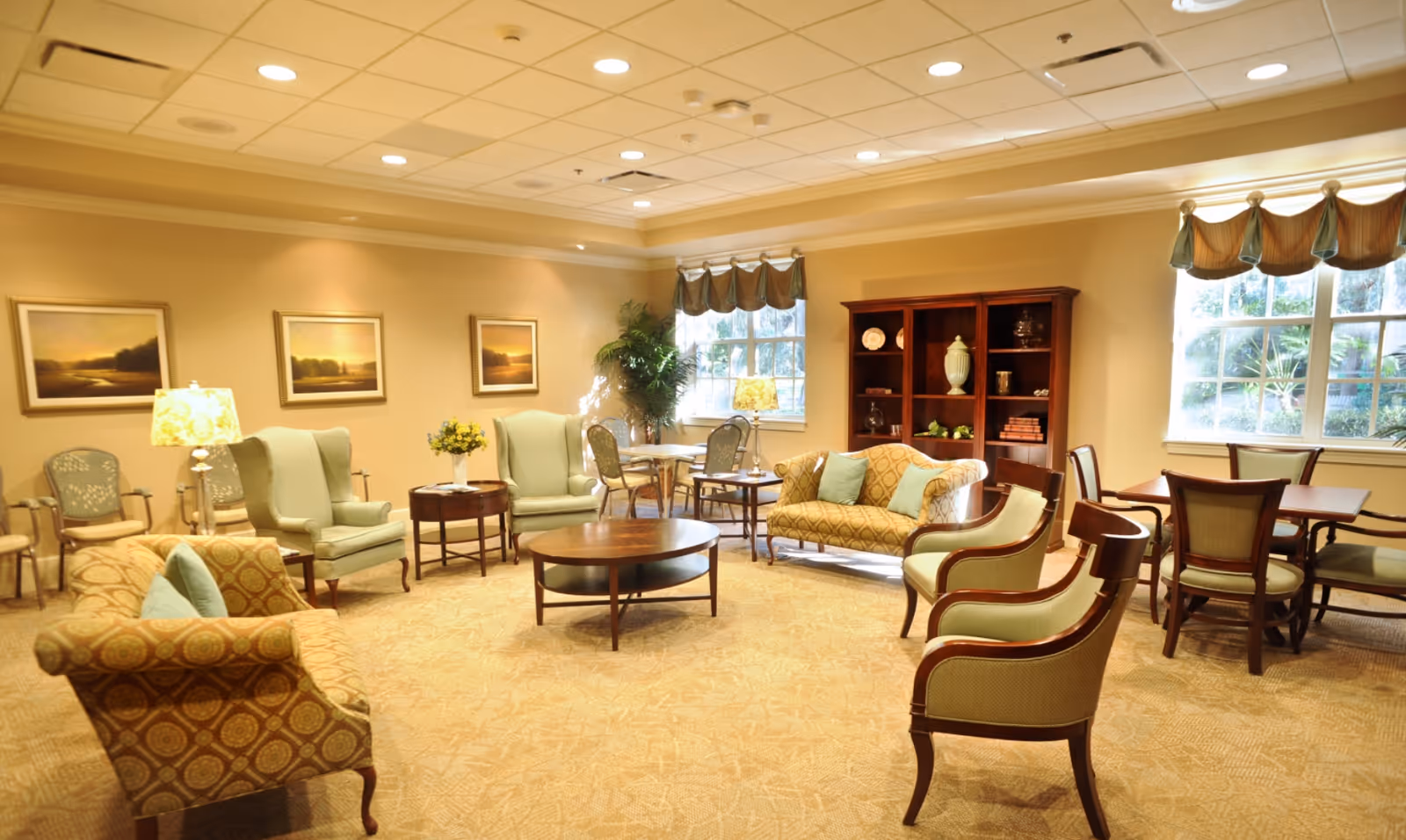 A well-lit senior living facility common area with comfortable seating including armchairs and sofas arranged around a central wooden coffee table. There are additional tables and chairs near windows with valance curtains, a wooden bookshelf with decorative items, framed landscape paintings on the wall, and a potted plant in the corner.