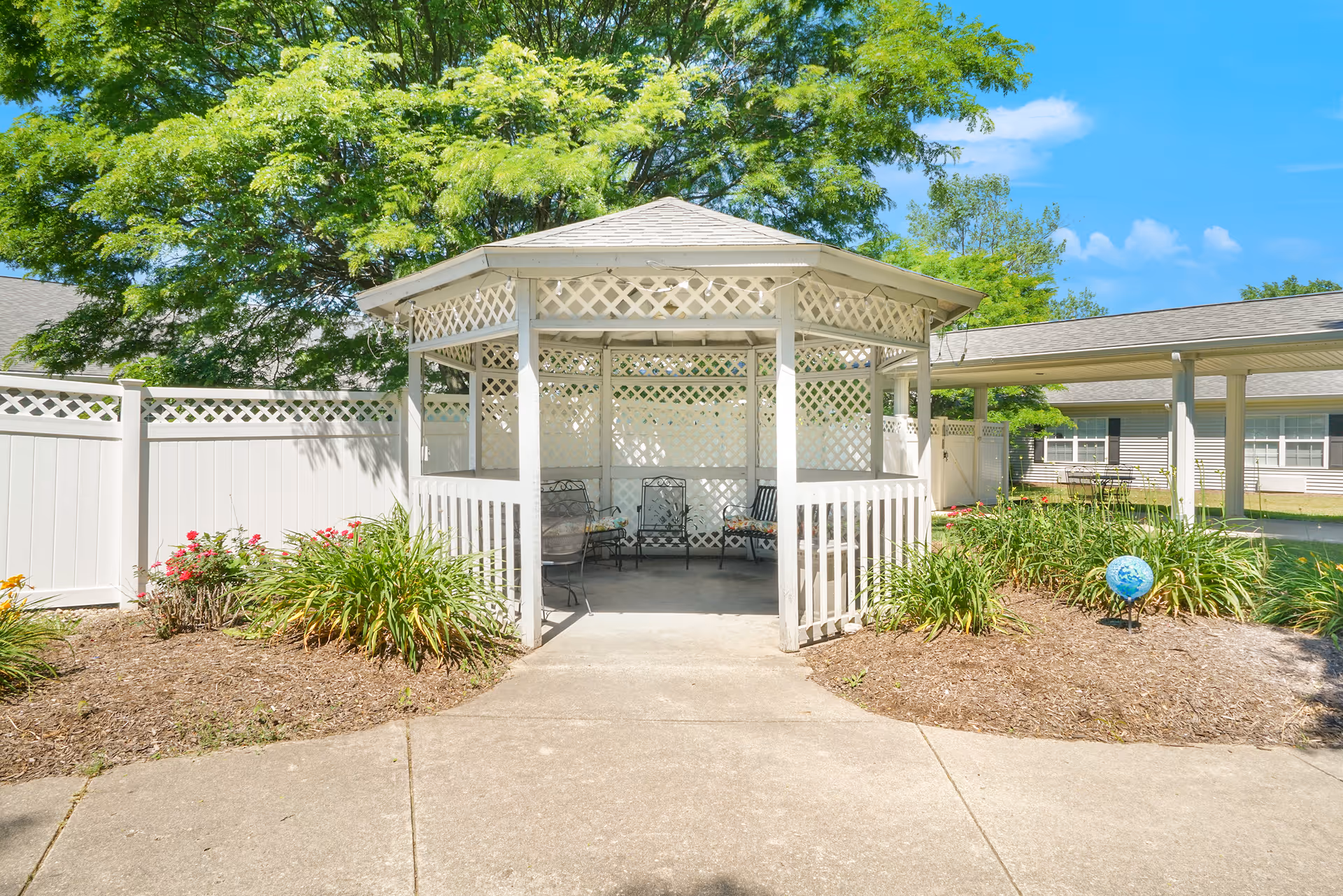 A white wooden gazebo with lattice panels and a shingled roof, surrounded by landscaped garden beds with green plants and flowers. The gazebo contains metal chairs and benches. In the background, there is a white fence, trees, and a building with windows under a clear blue sky.