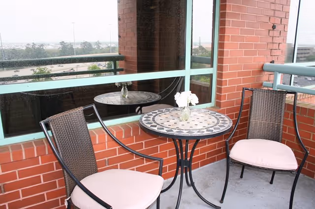 Small outdoor balcony area with two cushioned chairs and a round mosaic table with a small vase of white flowers, surrounded by red brick walls and a green railing, overlooking a distant view of trees and a parking lot.