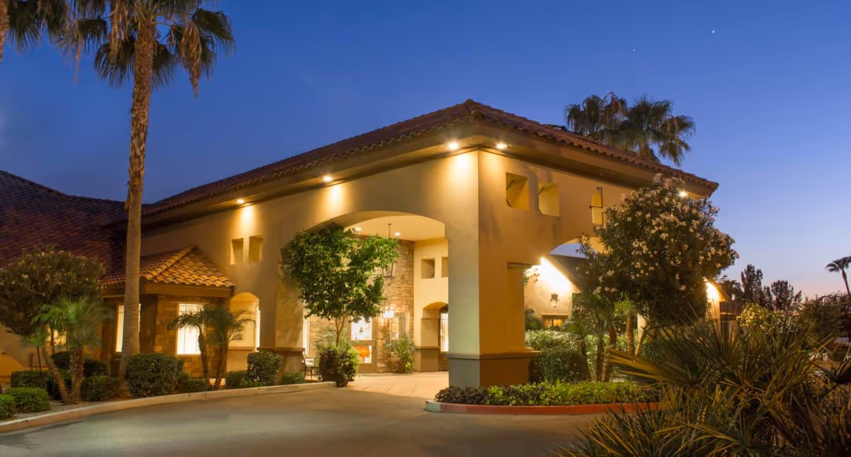 Exterior view of a senior living facility entrance at dusk with warm lighting, palm trees, and landscaped greenery surrounding the driveway and building.