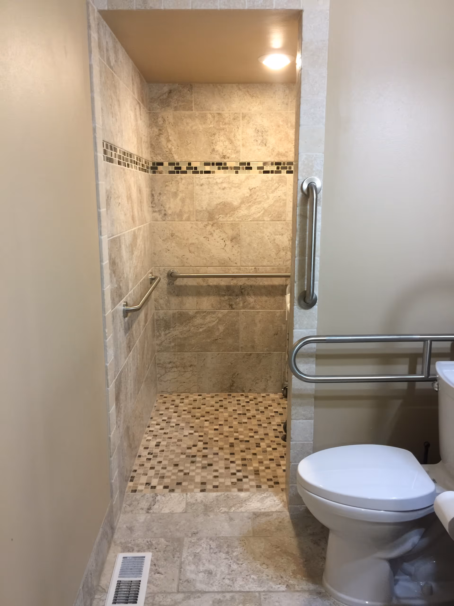 A bathroom with a tiled walk-in shower featuring a mosaic tile floor and beige wall tiles with a decorative horizontal strip. The shower has two metal grab bars installed on the walls. To the right, there is a white toilet with a metal grab bar next to it. The bathroom floor is tiled in a similar beige tone, and there is a floor vent visible near the entrance.