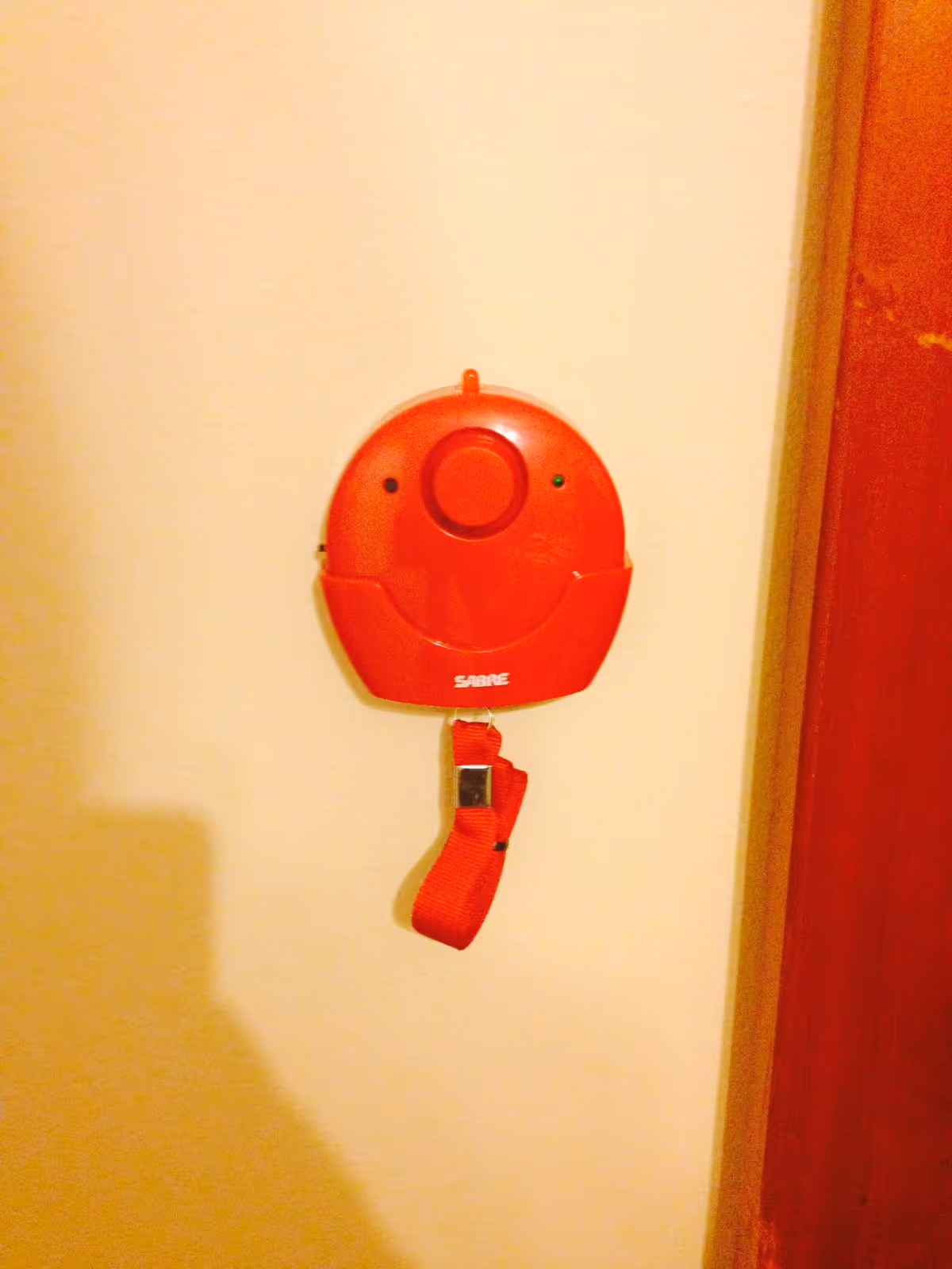 A red personal emergency alarm device with a wrist strap hanging on a beige wall next to a wooden door frame.
