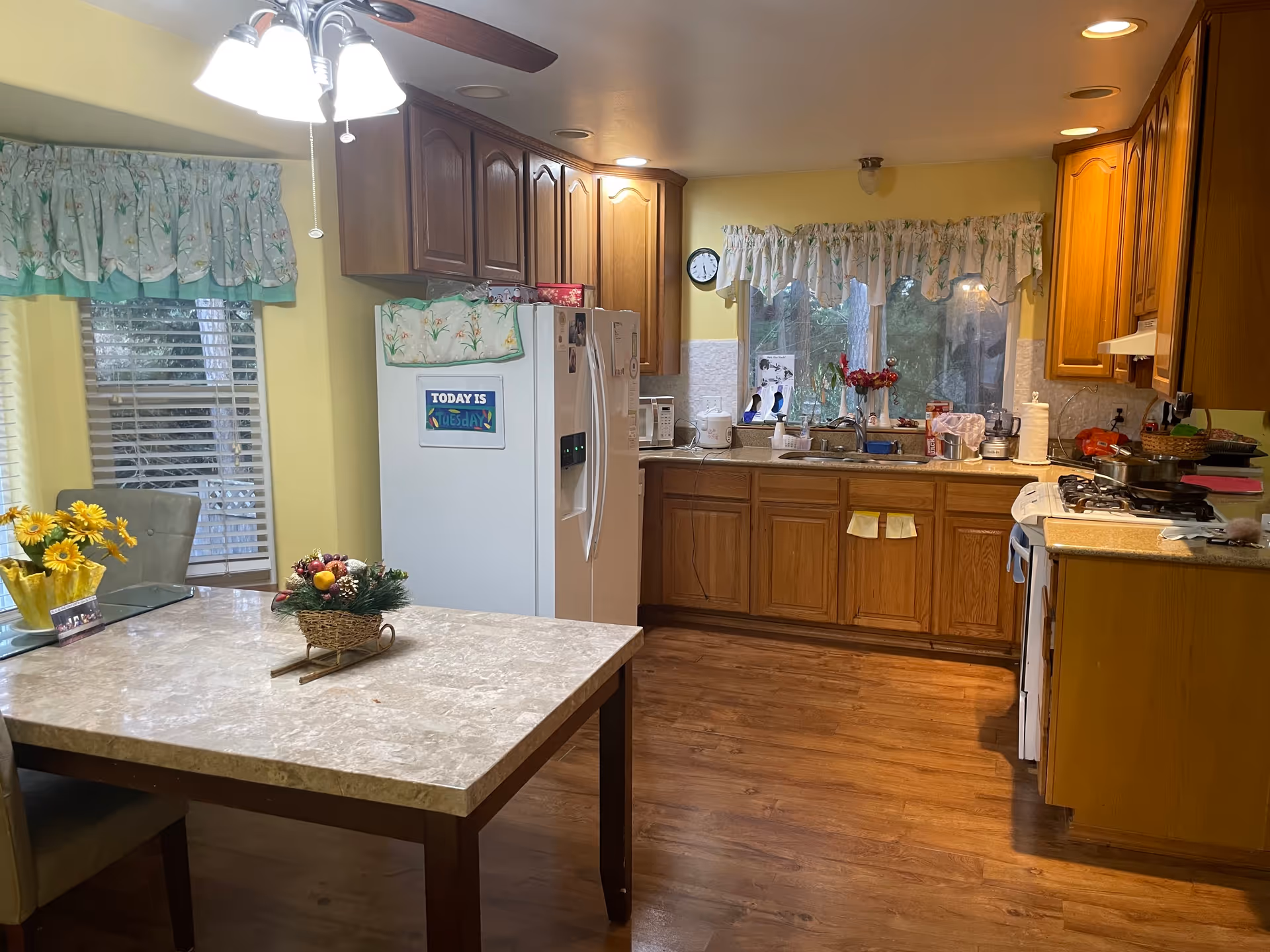 A cozy kitchen with wooden cabinets, a white refrigerator with a sign that says 'Today is Tuesday', a stove, and a sink under a window with floral curtains. There is a table with a marble-like surface and a flower arrangement in the foreground, and a ceiling light fixture above the table. The walls are painted yellow and the floor is wooden.