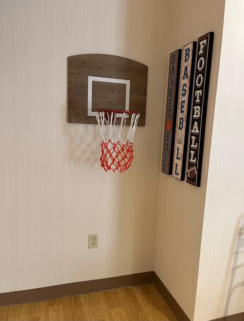 A small indoor basketball hoop mounted on a beige wall above a wooden floor. Next to the hoop are three vertical wooden plaques with the words BASKETBALL, BASEBALL, and FOOTBALL written on them, each with corresponding sports icons.