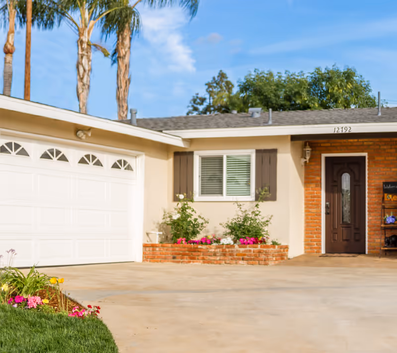 Front exterior view of a single-story house with a white garage door, a brown front door with a glass panel, brick accents around the entrance, a flower bed with colorful flowers, and palm trees in the background under a blue sky.