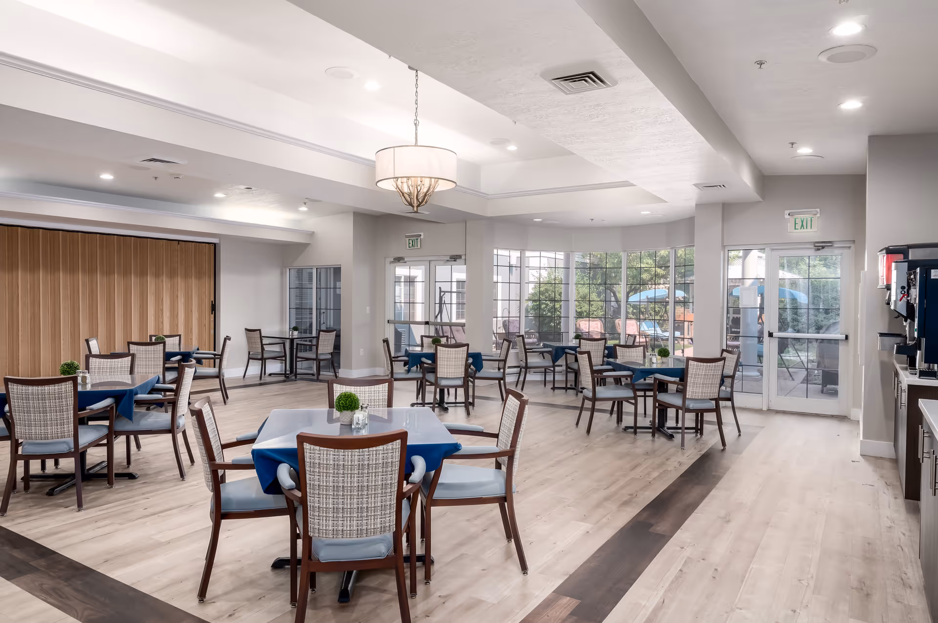 A bright and spacious dining room in a senior living facility with multiple tables covered with blue tablecloths and surrounded by wooden chairs with woven backs. Large windows let in natural light and offer a view of an outdoor patio area. The room features light wood flooring, a modern chandelier, and a beverage station on the right side near the exit door.