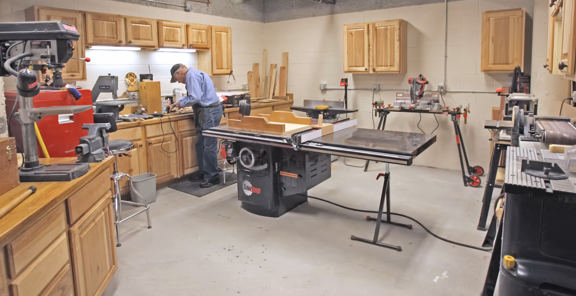 A woodworking workshop with various tools and equipment including a table saw, drill press, and workbenches with wooden cabinets. An elderly man wearing a cap and glasses is working at one of the benches.