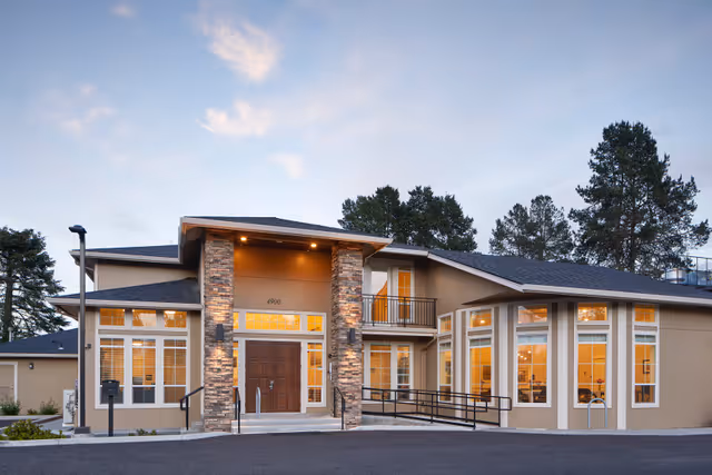 Exterior front of a modern memory care building featuring stone-clad columns, large windows, a central double-door entrance and an accessibility ramp at dusk.