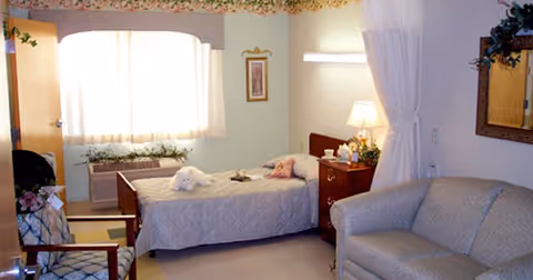 A cozy bedroom with a single bed covered in a light-colored quilt, a bedside table with a lamp and decorative items, a cushioned armchair, a sofa, a window with sheer curtains allowing natural light, and a framed picture on the wall.