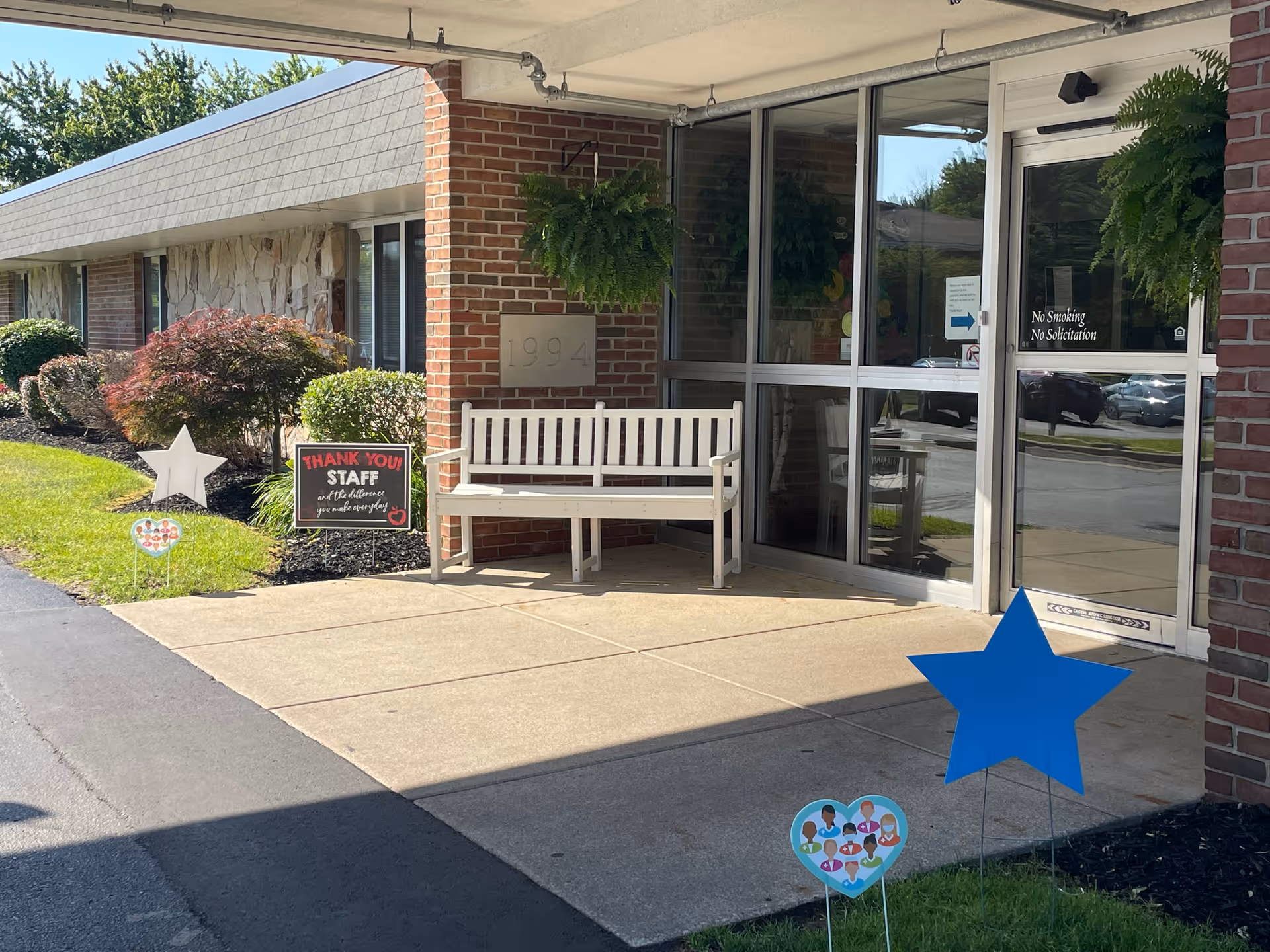 Entrance area of a facility with a white bench, two hanging green plants, and a glass door with a sign that reads 'No Smoking No Solicitation'. There are decorative signs on the grass including a blue star, a heart with diverse faces, and a sign thanking staff. The building has brick walls and the number 1994 is displayed on a plaque.