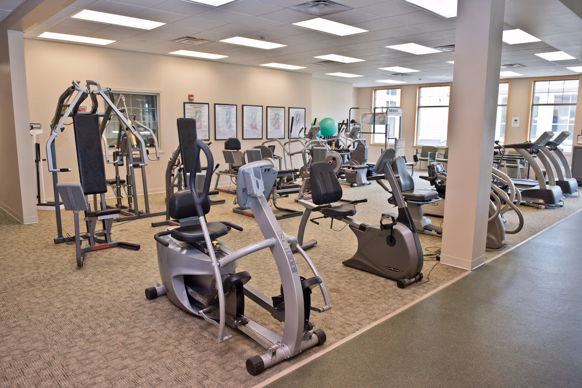 A well-lit fitness room with various exercise equipment including stationary bikes, treadmills, and weight machines. The room has large windows allowing natural light, beige walls, and carpeted flooring.