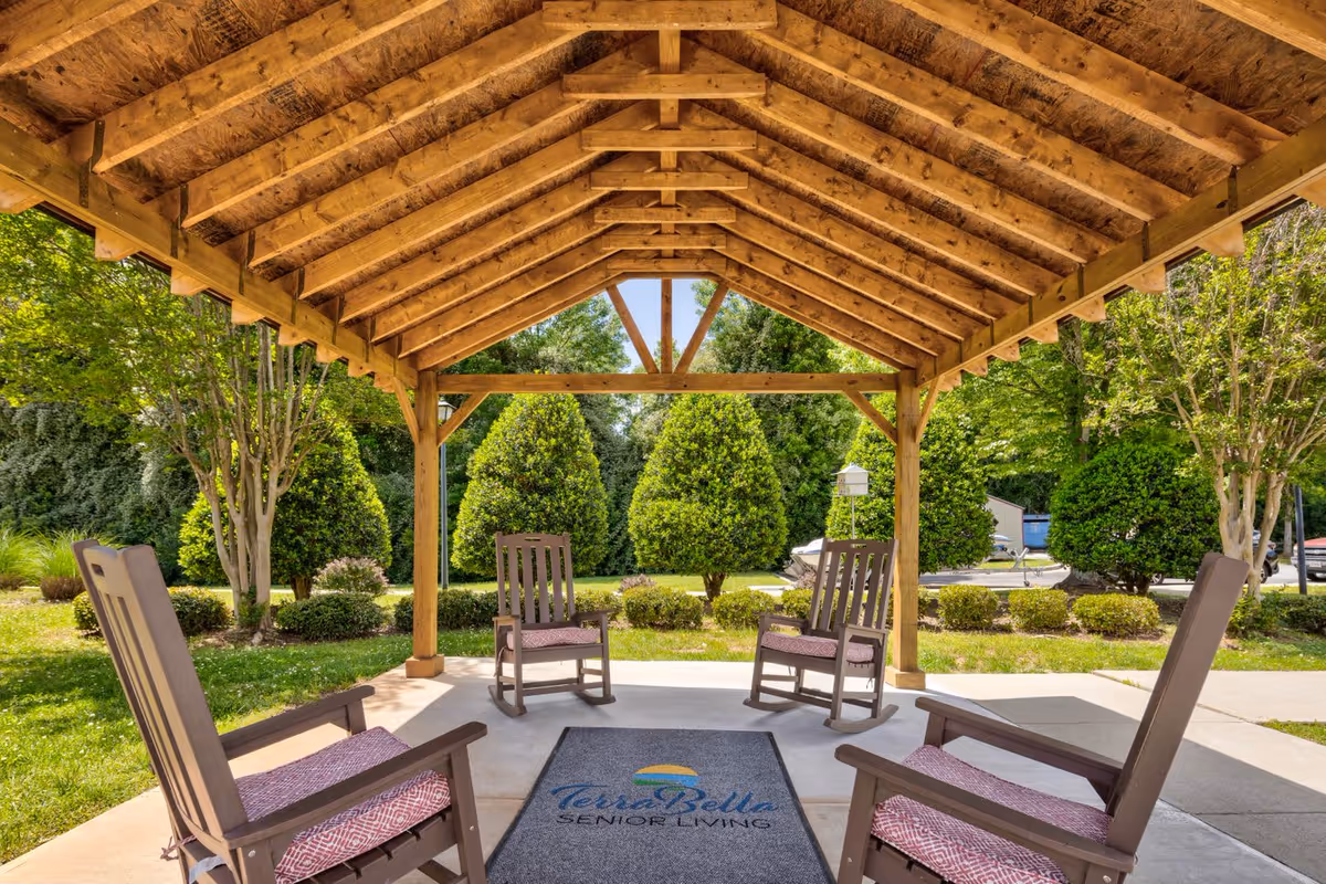 Outdoor covered seating area with a wooden roof structure and four wooden rocking chairs with cushions arranged around a TerraBella Senior Living branded mat, surrounded by green trees and bushes in a garden setting.