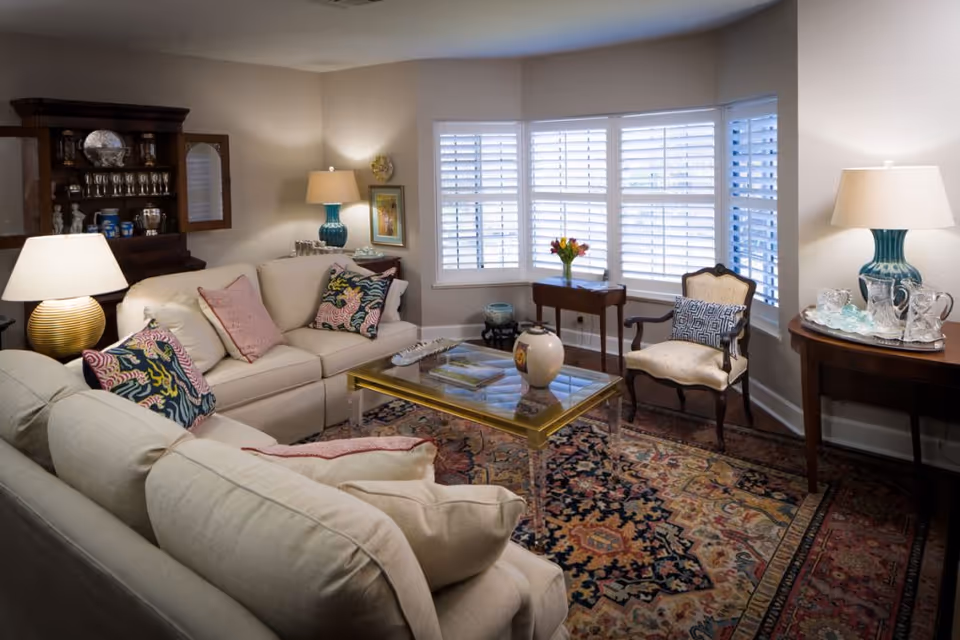 A cozy living room with beige sofas adorned with colorful patterned pillows, a glass coffee table with a decorative vase, a wooden armchair with a patterned cushion, and a side table with a lamp and crystal glassware. The room features a large window with white plantation shutters and a patterned area rug covering the floor.
