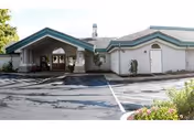 Exterior view of a single-story building with a covered entrance and a parking area in front. The building has a light-colored facade with green trim and a small garden area on the right side.
