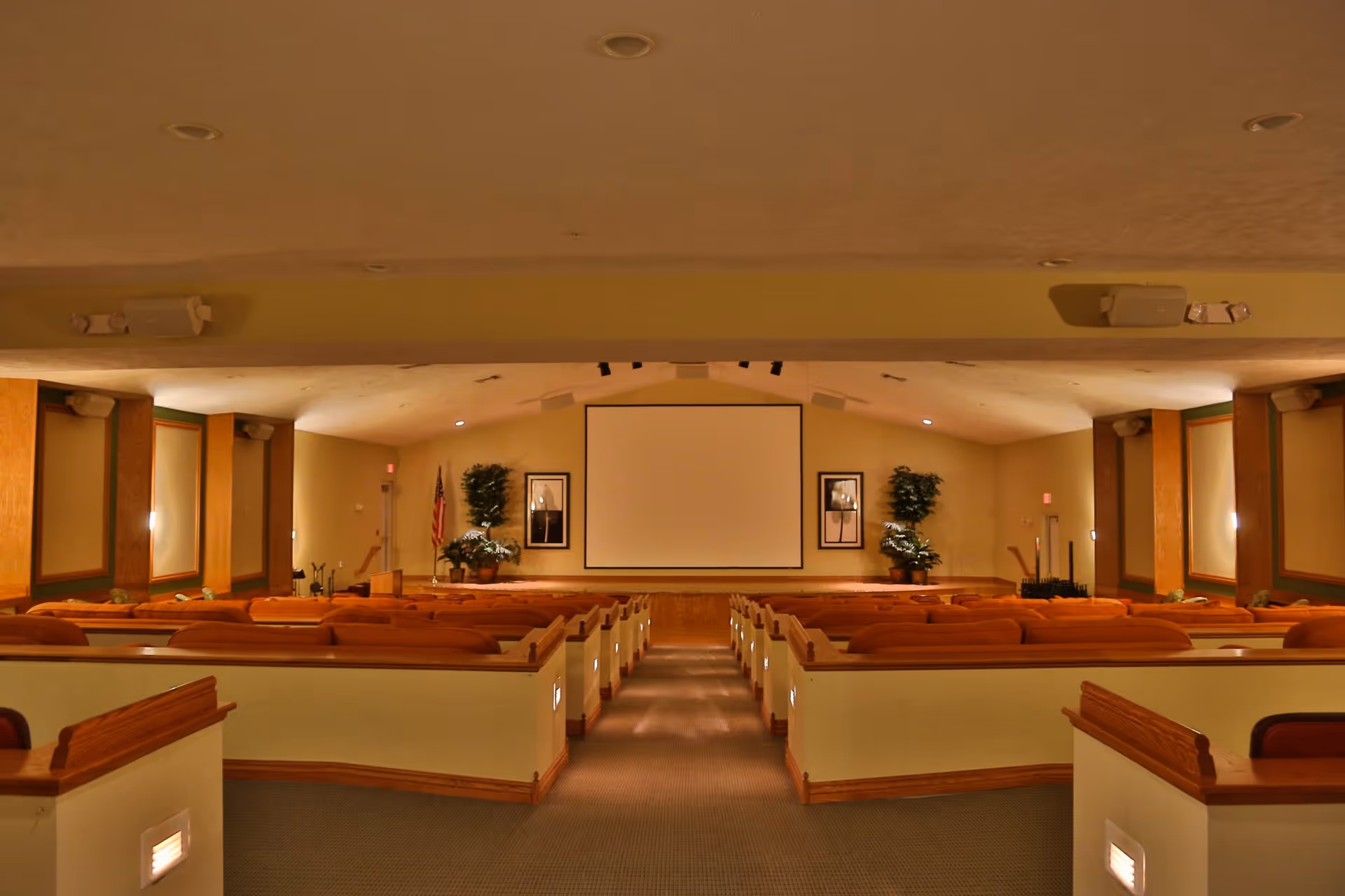 Interior view of a large auditorium or meeting room with rows of cushioned seats facing a stage with a large blank projection screen. The room has beige walls, framed pictures, potted plants, and an American flag near the stage.